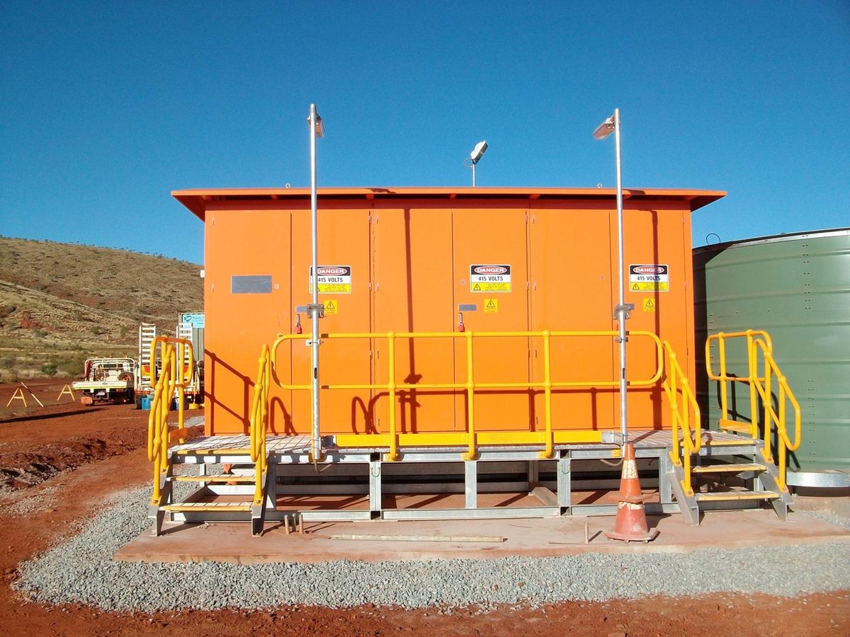 An orange plant switchroom on a concrete plinth with access stairs and handrails, a green storage tank alongside and hills beyond.