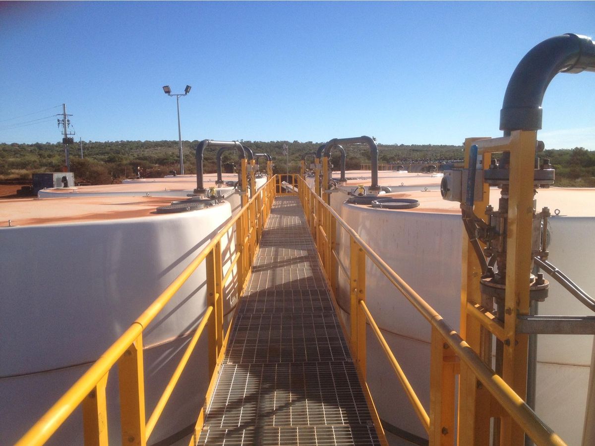 A grated walkway running between the tops of two treatment tanks, yellow handrails either side and process pipework rising from each tank.